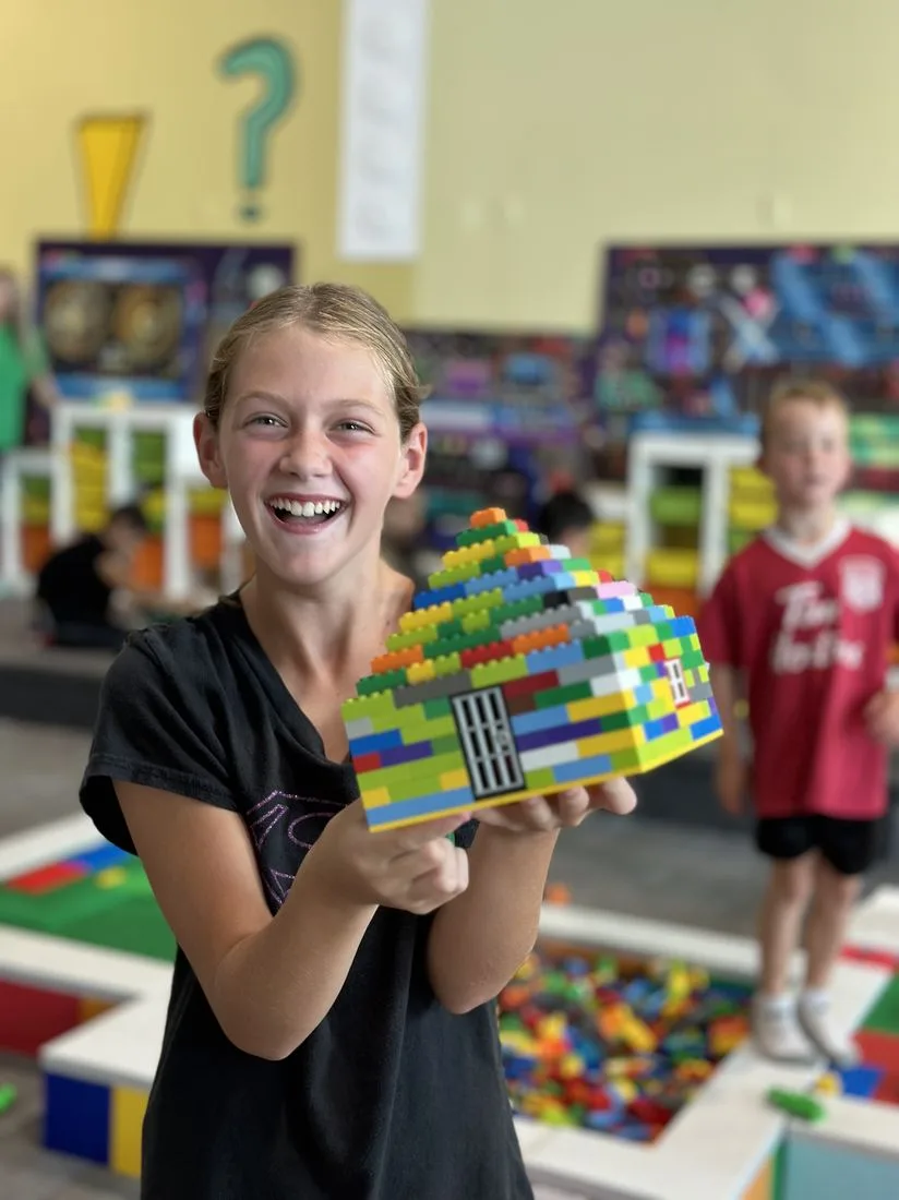 An older student laughing while holding up a large LEGO house she built
